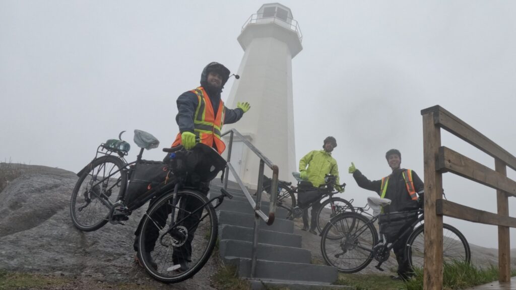 Three people stand with bikes in front of a lighthouse in the fog. 