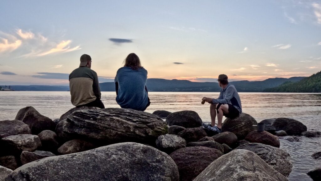 Three people sit on rocks along a lakeshore and look out toward a sunset. 