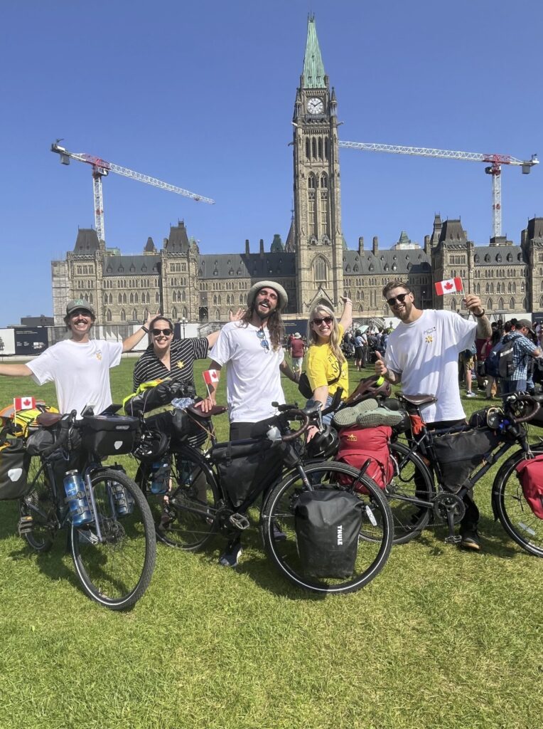 Five people with their arms outstretched next to three bikes stand in front of the government buildings and Canada flags. 