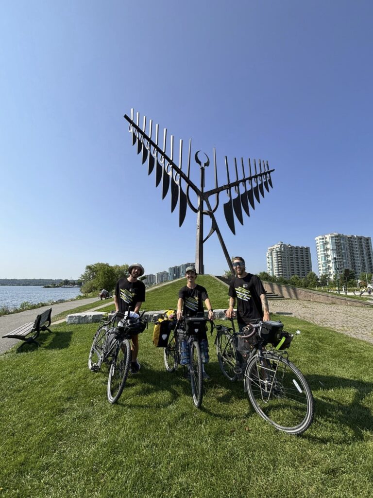 Three people with bikes stand in front of a large sculpture along a waterfront. 