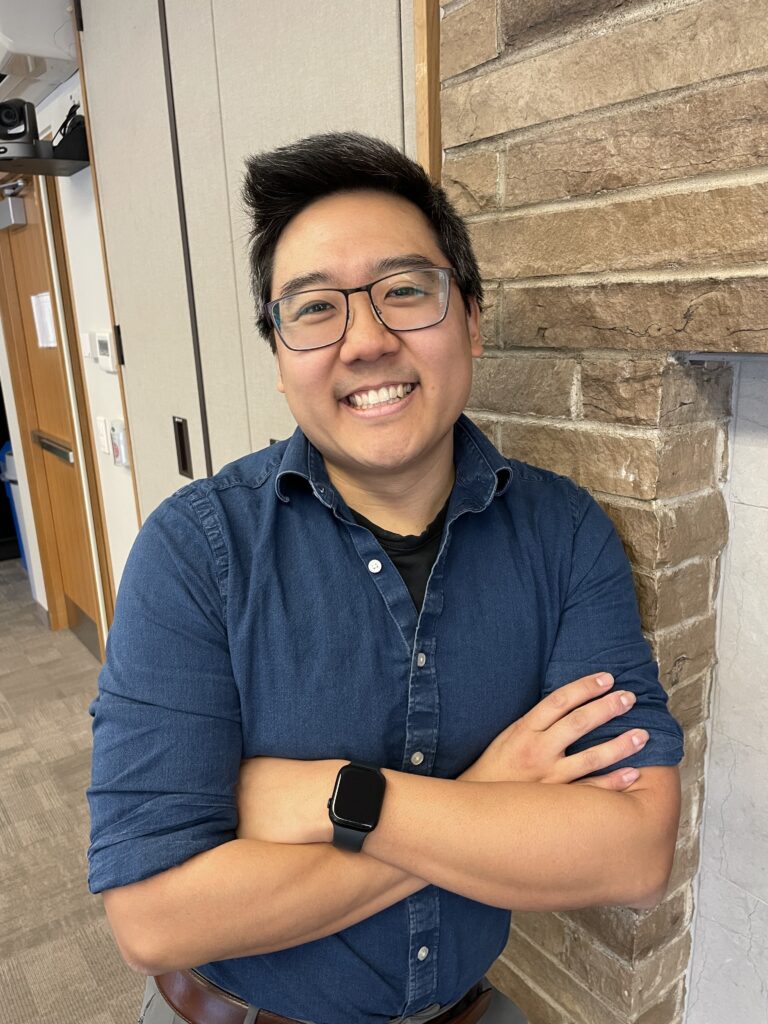 A person smiles with their arms crossed against their chest and leans against a wall.