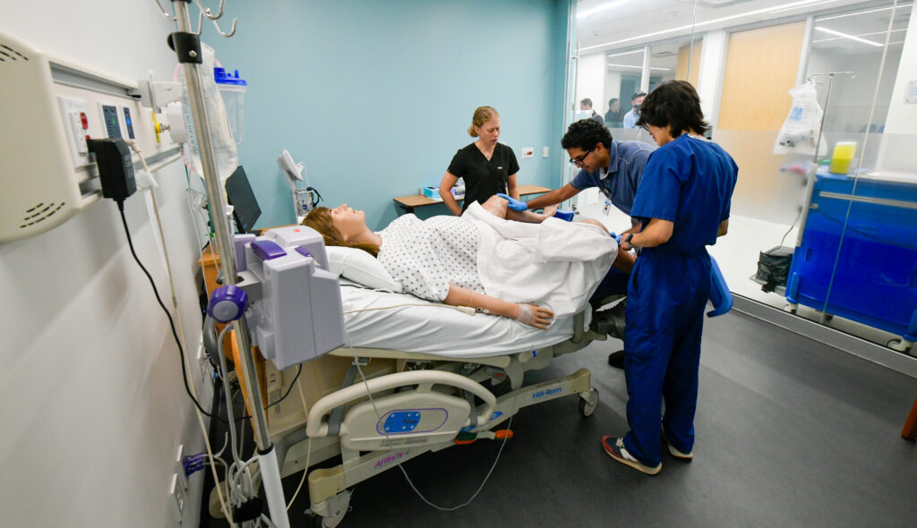 Three people stand around the legs of a high-fidelity manikin, lying on a hospital bed, that is simulating a postpartum hemorrhage. 