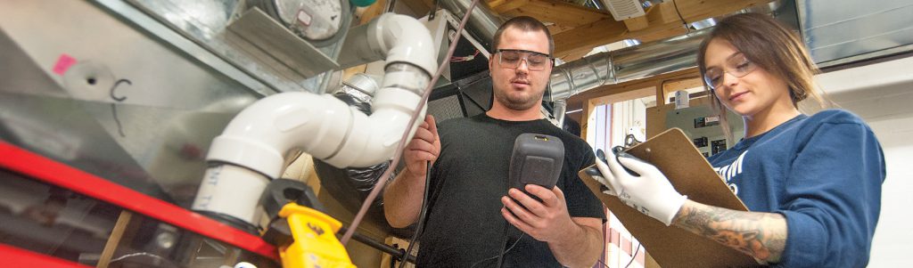Two HVAC technicians wearing safety glasses and holding a multimeter and clipboard while assessing a piece of equipment