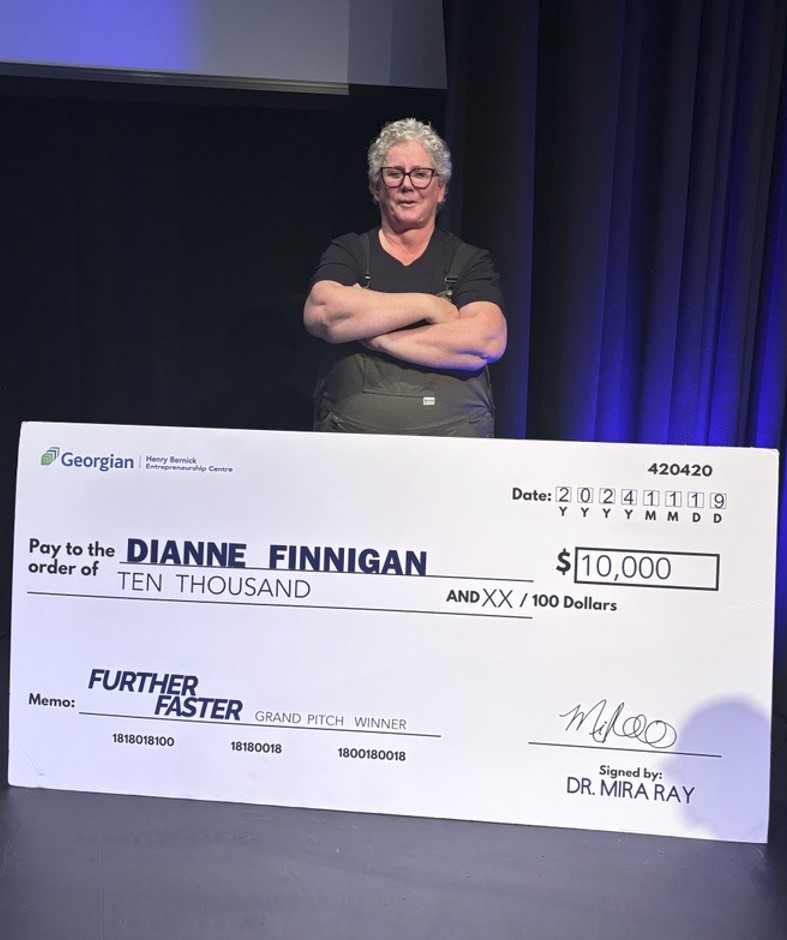 A person stands with their arms crossed on a stage behind an oversized cheque for $10,000.