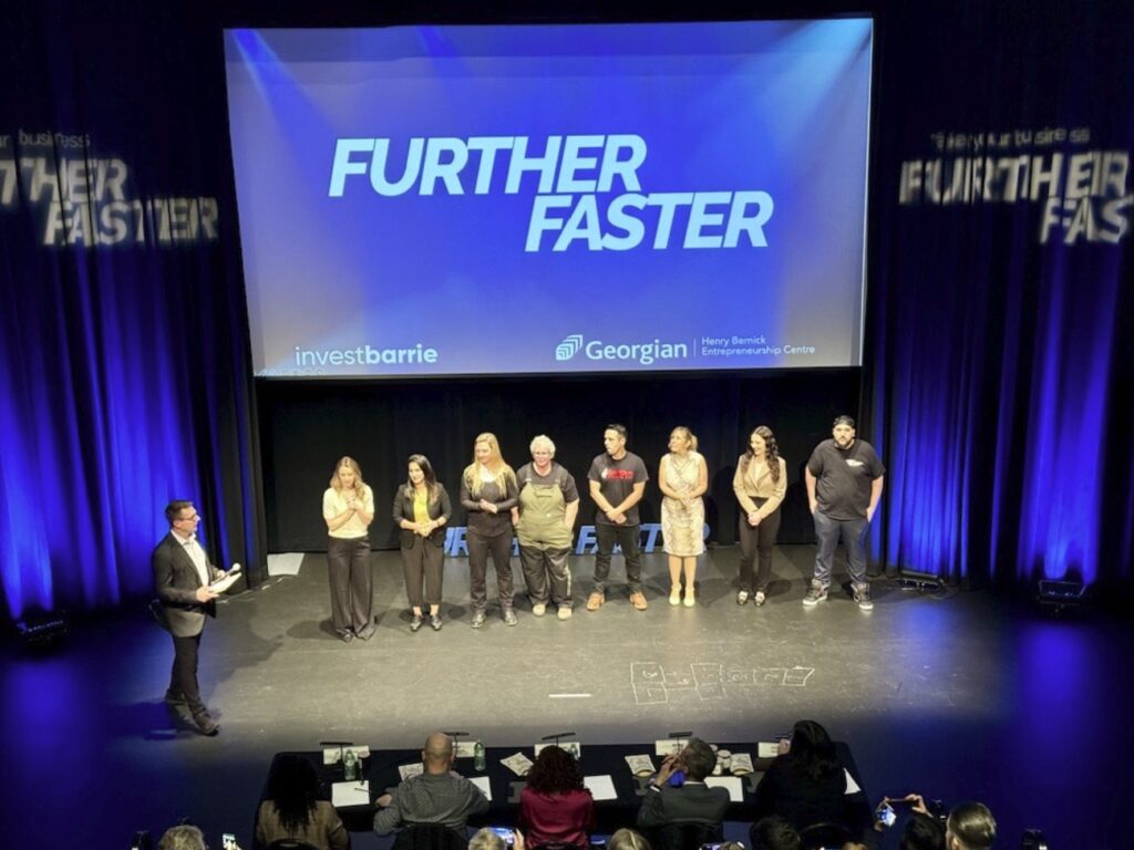 Eight people stand in a row on a stage, with another person standing nearby holding a microphone. Signage behind them reads: Further Faster.