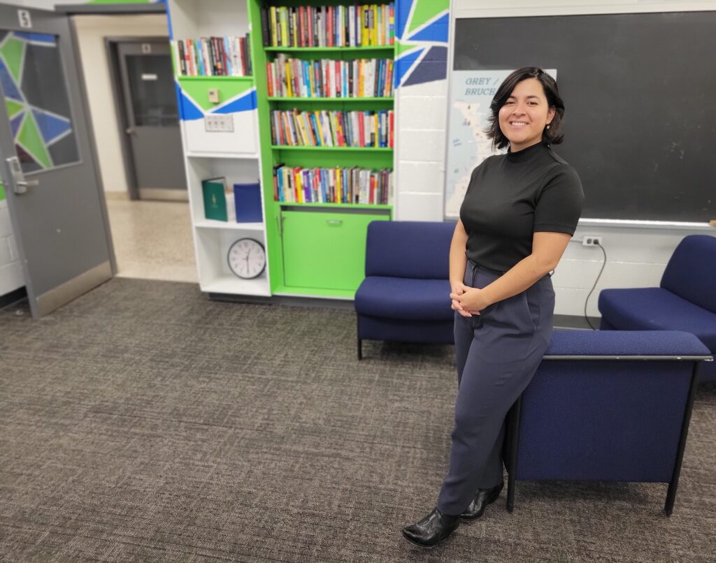 A person stands in a room with chairs, bookshelf, blackboard and more. 