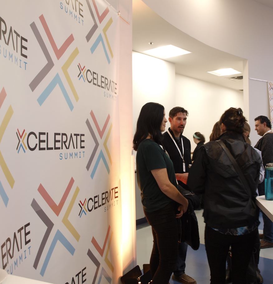 A group of people talk to each other next to a backdrop reading Xcelerate Summit.