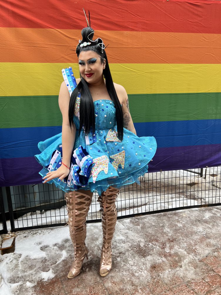 A drag queen standing outside in front of a rainbow flag, and wearing a blue dress and gold boots.
