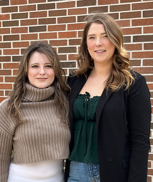 Katie Cannell and Jessica Davies-Thompson, graduates of the Honours Bachelor of Interior Design (BAID) program at СŶƵ, standing in front of a red brick wall smiling and looking at the camera