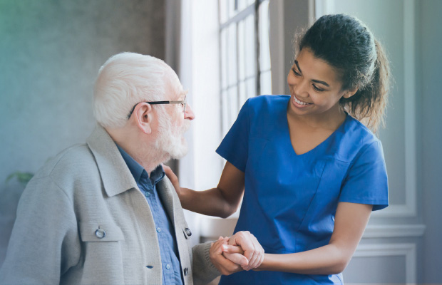 A Registered Nurse in blue scrubs smiling and assisting an elderly patient with getting up