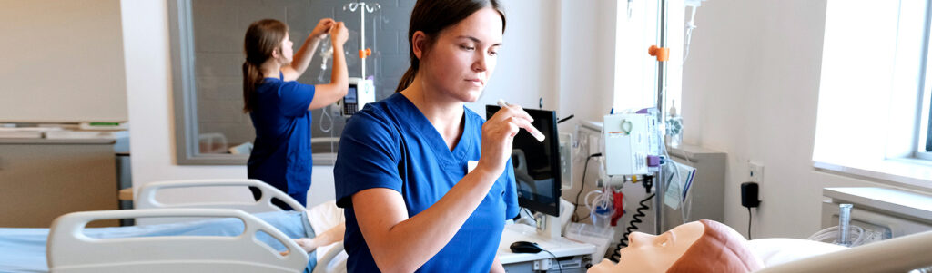 BScN nursing student in blue scrubs checking on a human patient simulator in the nursing lab.