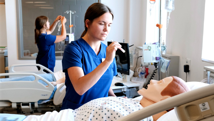 A woman in blue scrubs analyzes a manekin in a hospital room. 