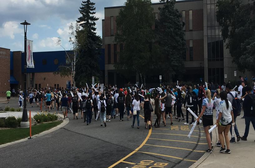 A large group of college students walk across campus in front of buildings.