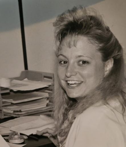 A black-and-white photo of an adult sitting at a desk in front of a pile of papers and turning toward the camera with a smile.