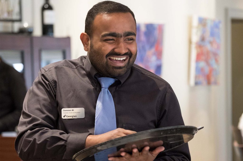 Hospitality, Tourism and Recreation student wearing a black collared shirt, blue tie and name tag while holding a tray in the Georgian Dining Room