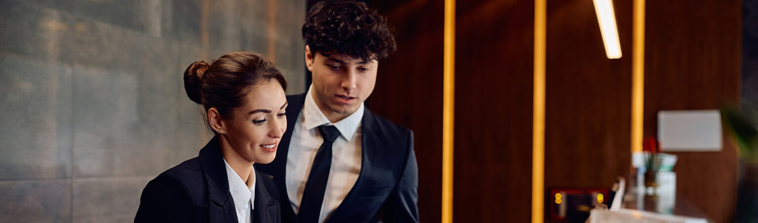 Two hospitality professionals in formal business attire stand at a modern hotel reception desk, reviewing information together in a warmly lit lobby.