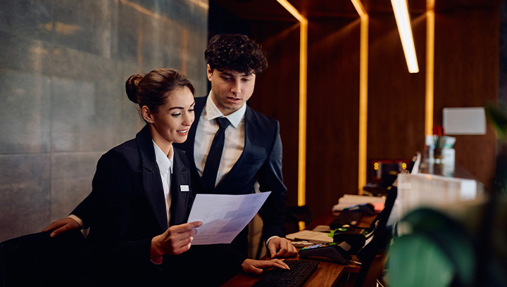 Two hospitality professionals in formal business attire stand at a modern hotel reception desk, reviewing information together in a warmly lit lobby.