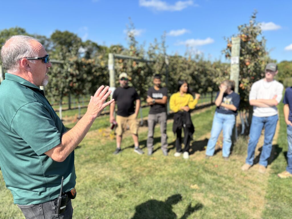 A person stands outside in an apple orchard in front of a group of people. 