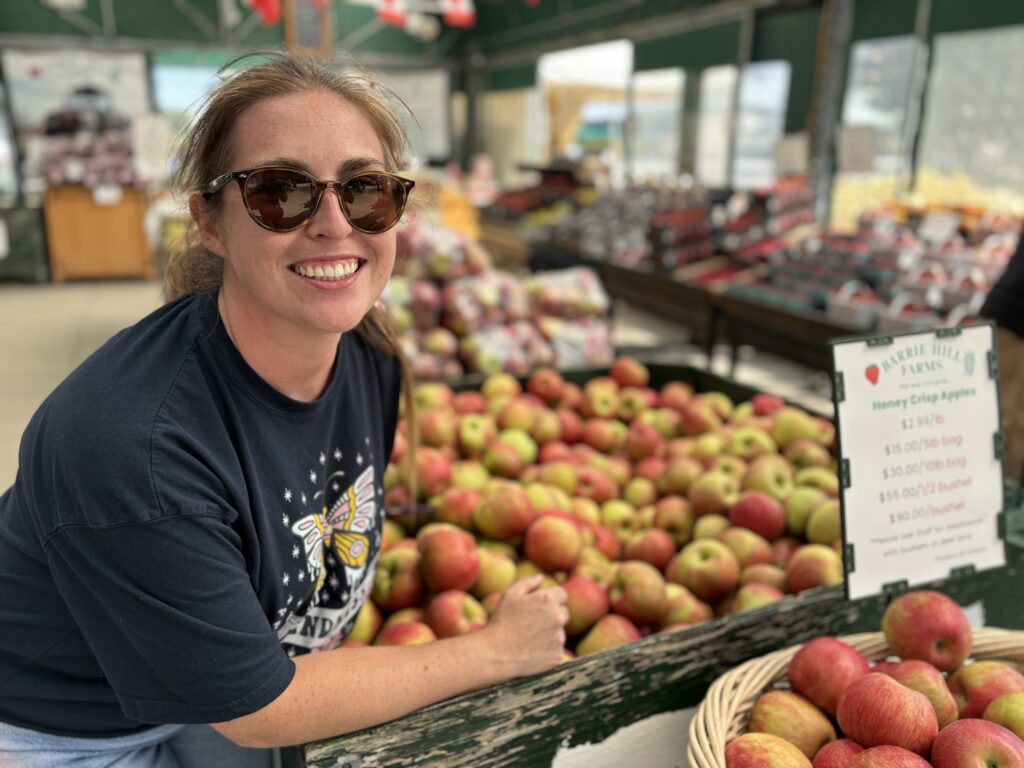 A person stands in a fruit and vegetable market next to a large box of apples. 