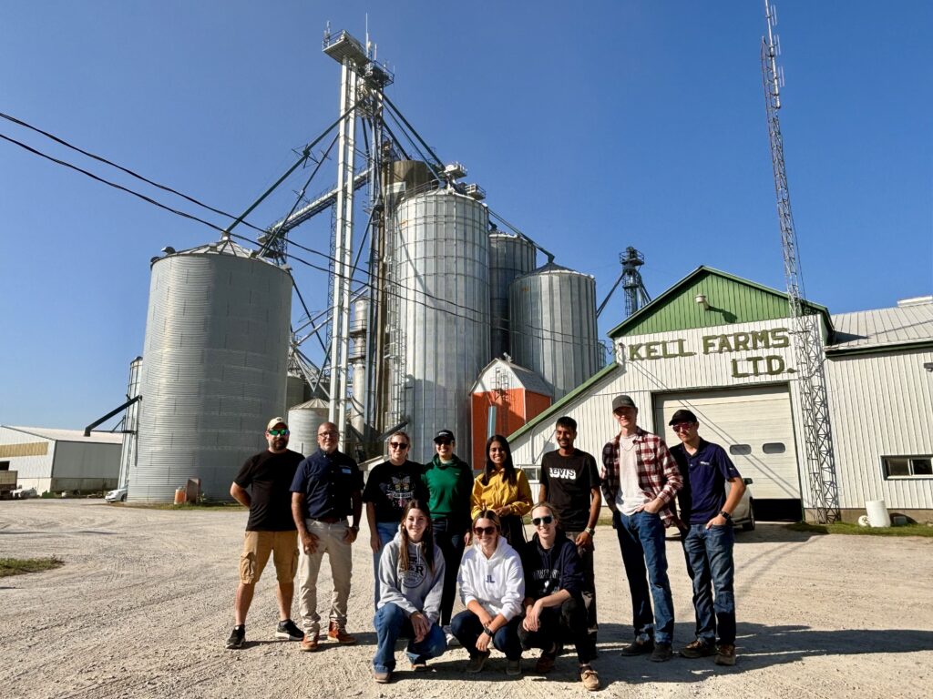 A group of people stand outside in front of grain elevators. Sign on a building reads: Kell Farms Ltd.