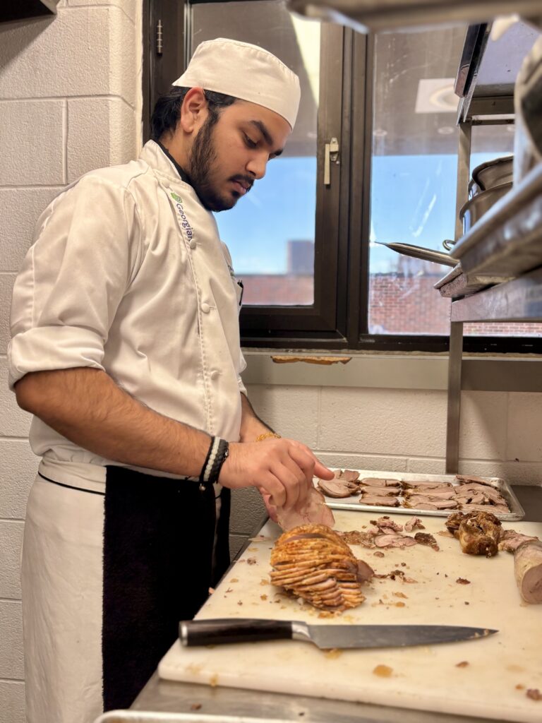 A person wearing a chief's uniform and hat stands in a kitchen and cuts slices of turkey. 