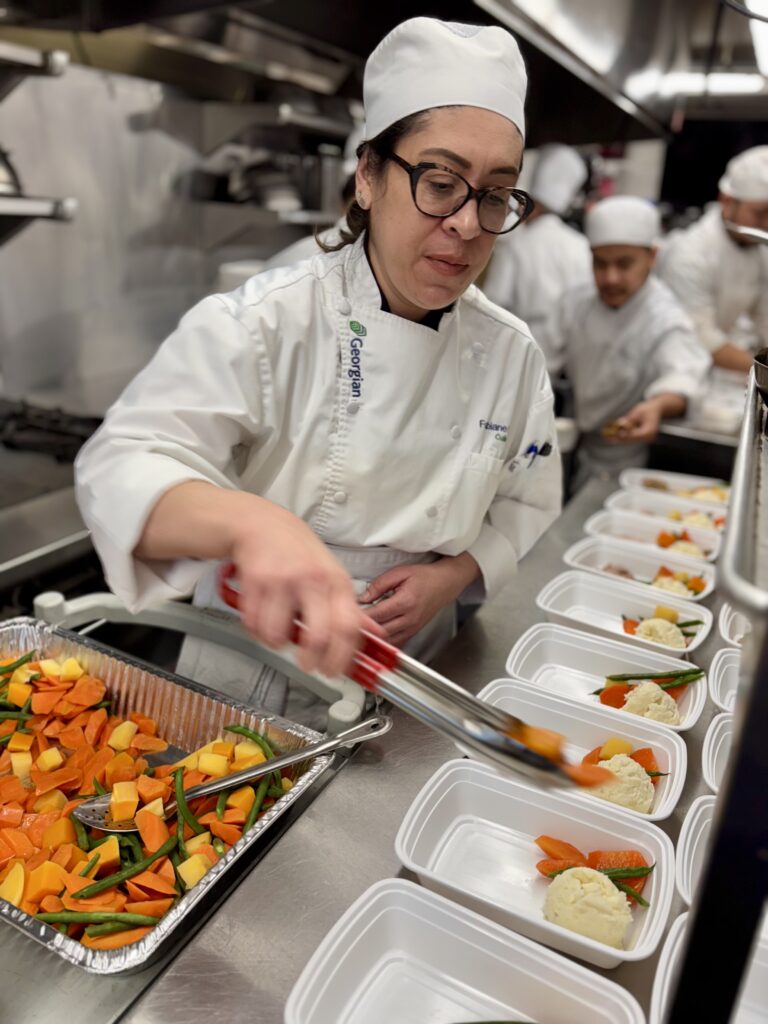 A person wearing a chef's uniform and hat in a kitchen dishes out vegetables into individual containers. 