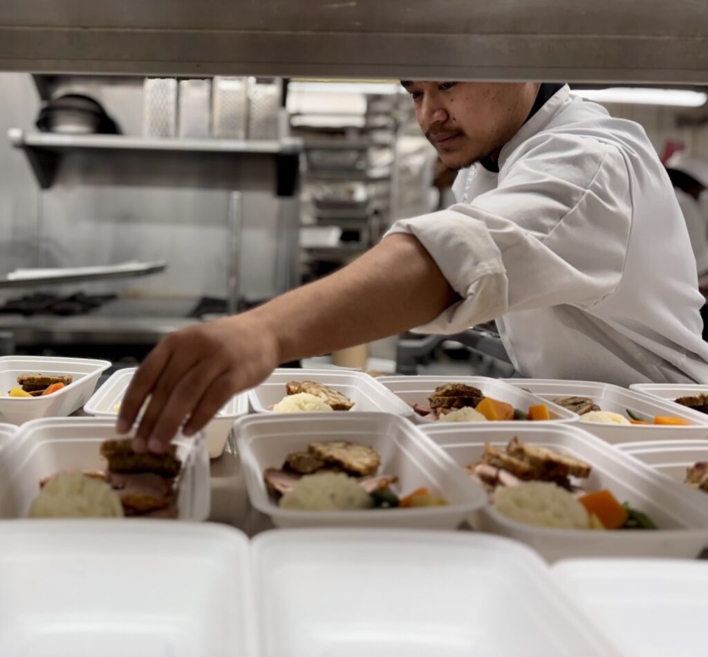 A person wearing a chief's uniform reaches out to add stuffing into individual meal containers. 