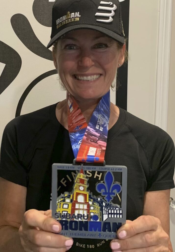 A person wearing a black T-shirt and hat wears a medal around their neck and holds it up. It reads "Subaru Ironman."