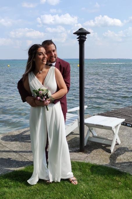 Two adults, wearing a suit and wedding dress, respectively, pose in front of a lake with their arms around each other.