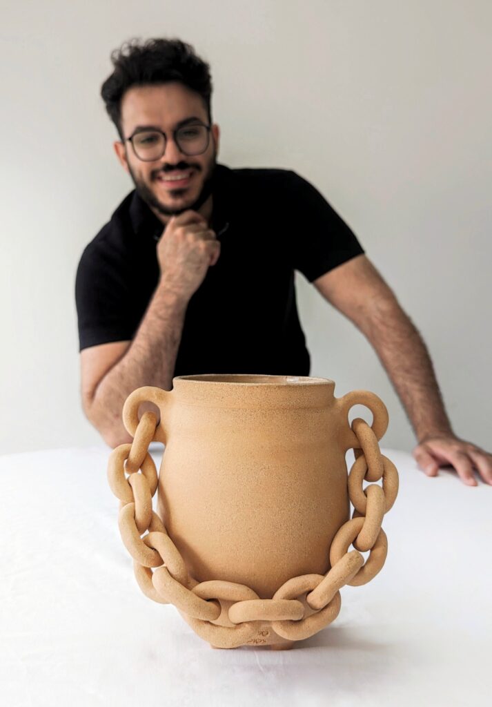 A person sits in the background with a beige decorative vase with a thick chain across the front sits in the foreground.