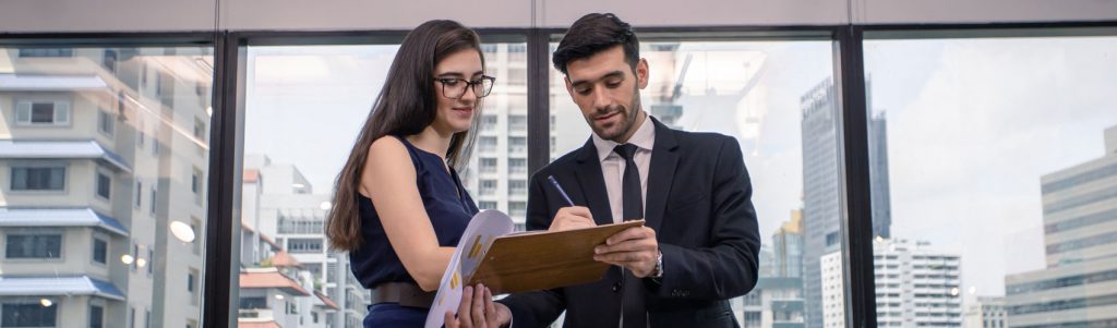 Person in a suit and tie holding out a clipboard with paperwork while a colleague wearing glasses and a dress signs in pen