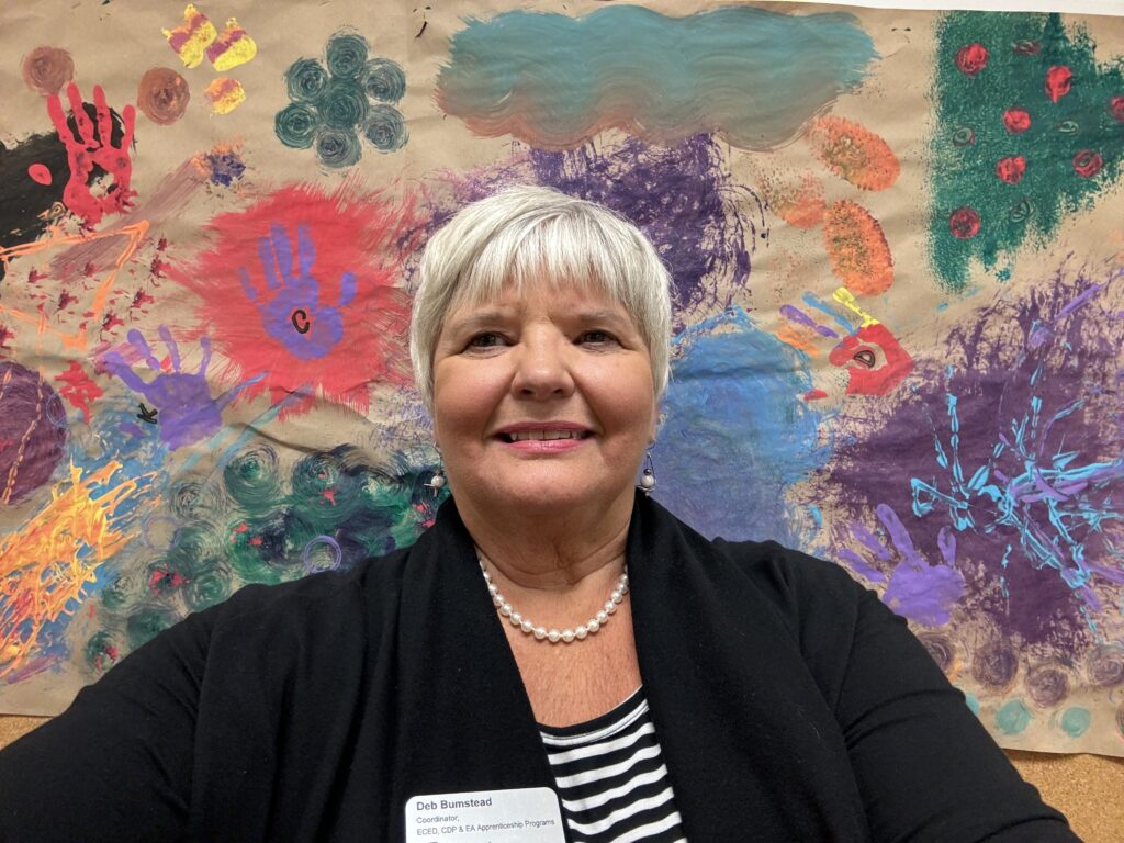 A person with short white hair and black sweater takes a selfie in front of a paper backdrop covered in kids' paintings.