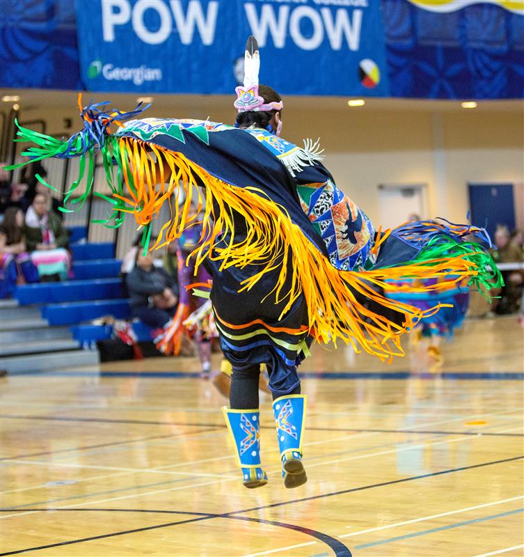 A person wearing Indigenous regalia dances in a gymnasium. A sign reads "Pow Wow."