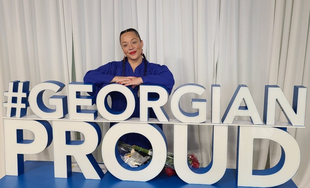 A person wearing a blue convocation robe stands behind a marquee sign reading #GeorgianProud.