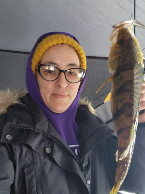 A person wearing a winter jacket and toque holds up a fish. 