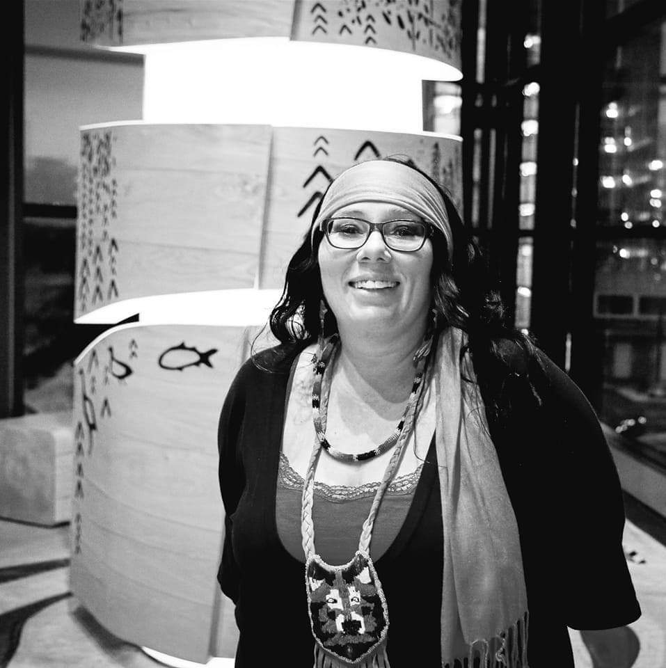 A black-and-white image of a person dressed casually with a scarf tied around their dark hair, smiling in front of Indigenous artwork they created.