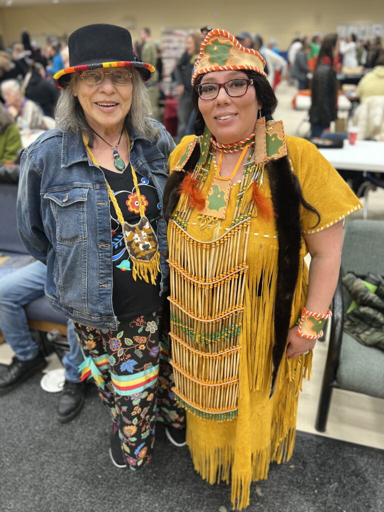 Two people stand together. One is dressed in a ribbon skirt, jean jacket and black, beaded hat, and the other is wearing a buckskin dress.