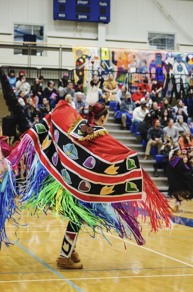 A person wearing colourful, Indigenous regalia dances in a gymnasium. 