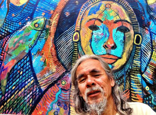 An older man (Paul Shilling) with long gray hair and a beard standing in front of a colourful piece of artwork.
