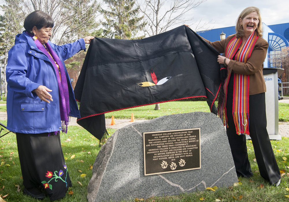 Elder Lorraine McRae and President and CEO MaryLynn West-Moynes unveil the Land Acknowledgment plaque on a stone outside at the Barrie Campus