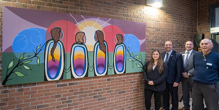 Four people standing near a brick wall with a piece of colourful artworkk on it. The four people are Emily Kewageshig, Kevin Weaver, Dave Shorey and Greg McGregor. They're all dressed in business attire.