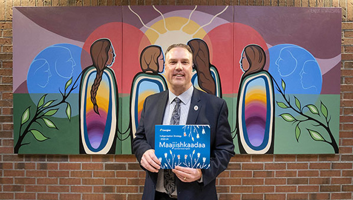 Presient Kevin Weaver dressed in business attire standing in front of a piece of Indigenous artwork. He's smiling and holding up a booklet.
