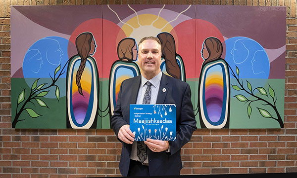 Presient Kevin Weaver dressed in business attire standing in front of a piece of Indigenous artwork. He's smiling and holding up a booklet.