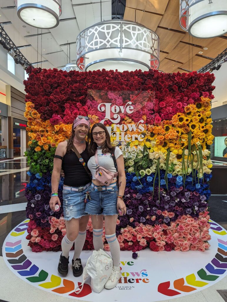 Two people stand together in front of a wall of flowers in rainbow colours. A sign reads: Love grows here.