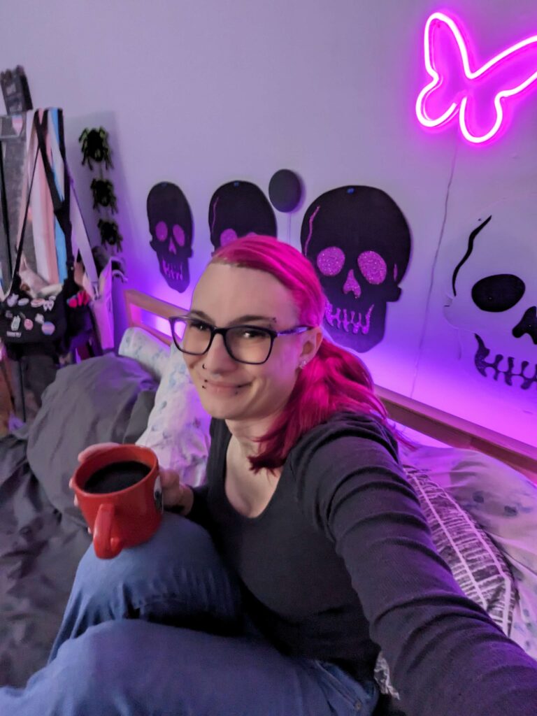 A person sits on a bed while holding a cup of coffee and takes a selfie.