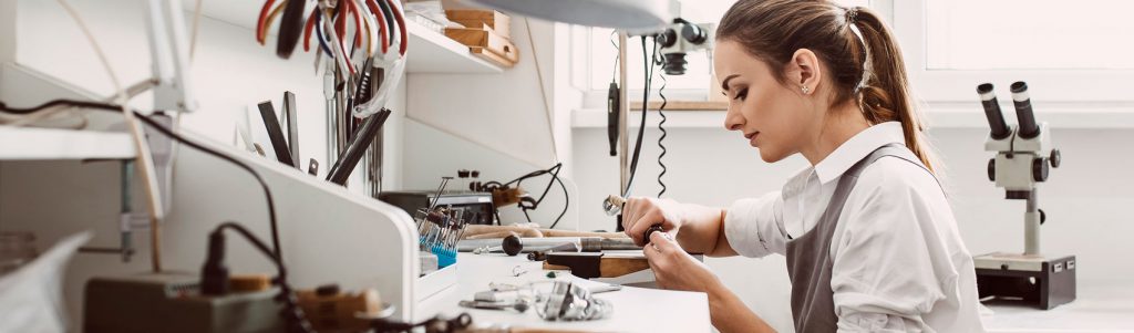 A jewellery maker wearing an apron while sitting at a workstation in the studio using tools to forge a piece of jewellery