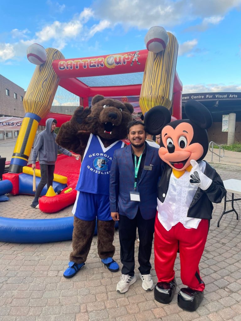 A person wearing a suit smiles in between a grizzly bear mascot and Mickey Mouse, standing in front of an inflatable carnival game.