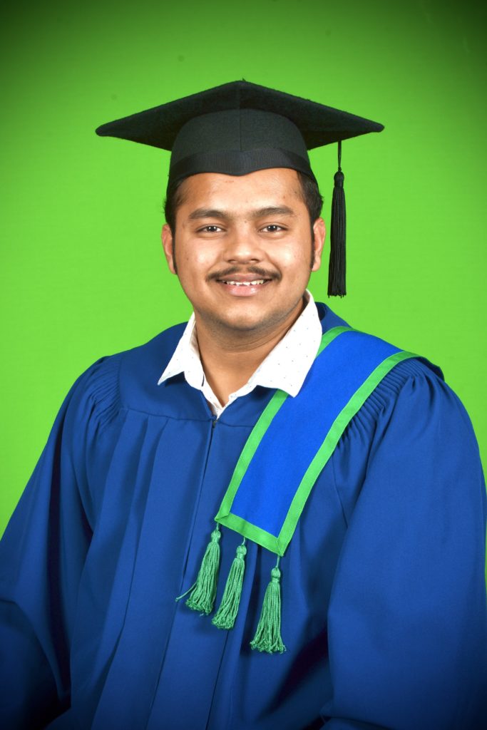 A person dressed in a blue graduation gown and cap smiles at the background.