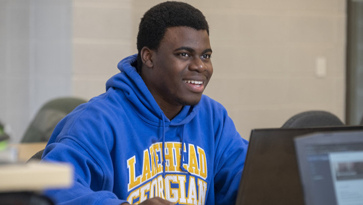 Male student wearing a Lakehead-Georgian blue sweatshirt while using a laptop computer and smiling
