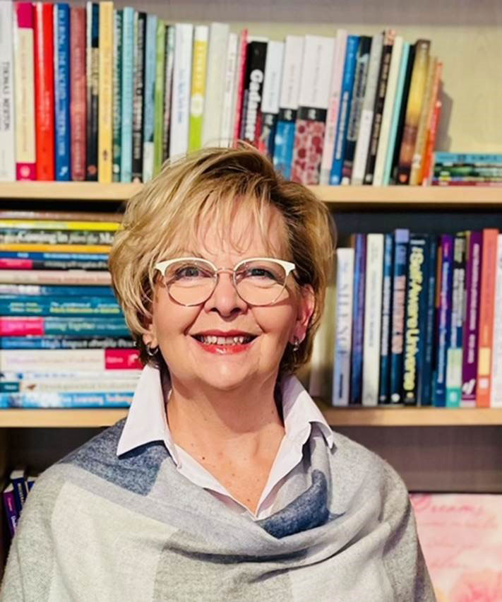 A smiling woman (Dr. Anne-Liisa Longmore) with short blonde hair and glasses standing in front of a bookcase